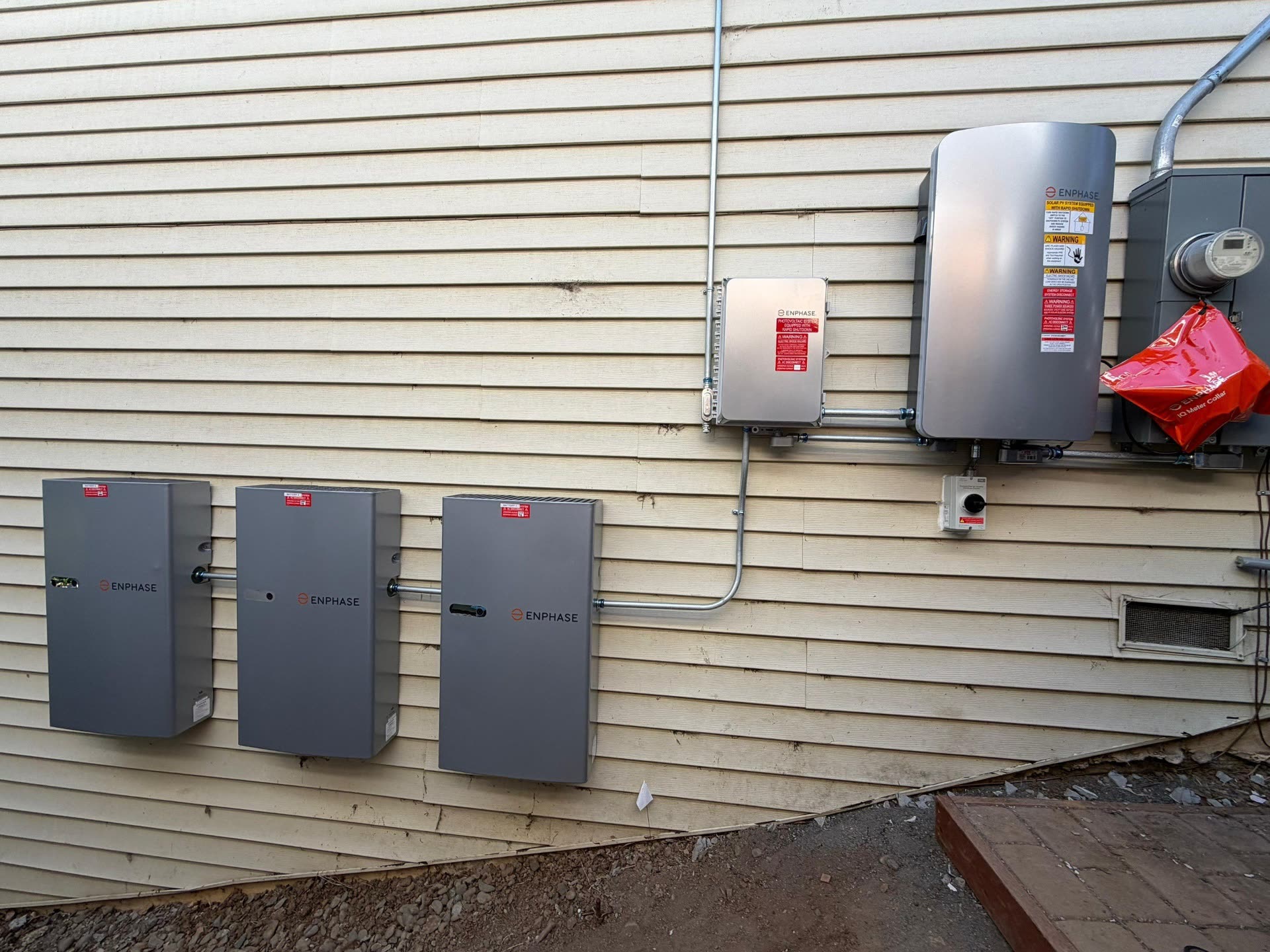 Three Enphase IQ Batteries mounted along the exterior wall of a Belmont home alongside the Enphase system controller and utility meter collar.