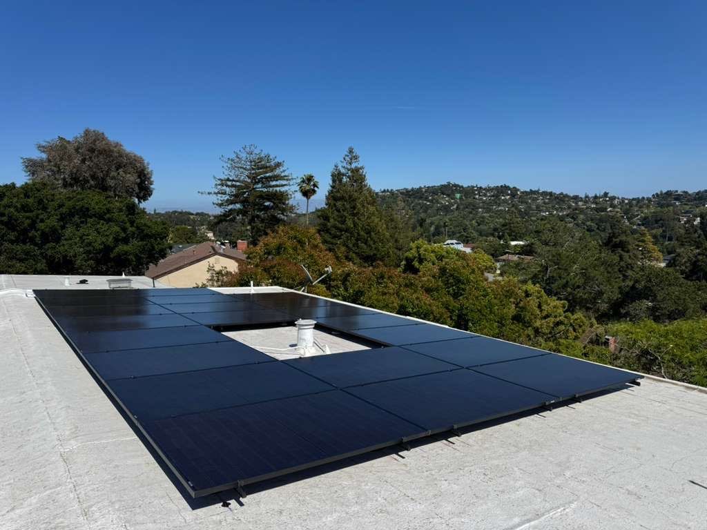 All-black solar array installed on a low-slope white roof of a Belmont home, overlooking tree-lined hills and neighborhoods.