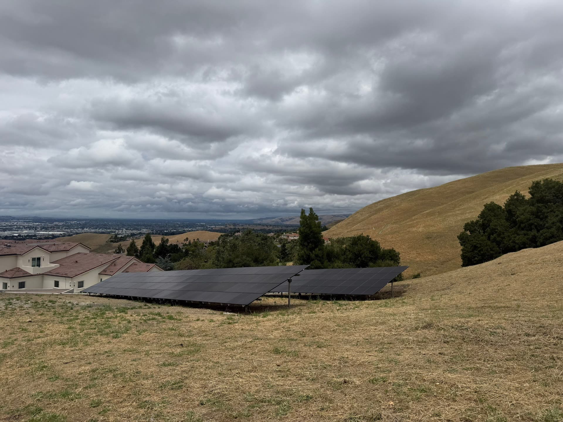 Residential ground-mount solar array of all-black panels installed on a hillside above Fremont, CA with the valley and rolling hills in the background.