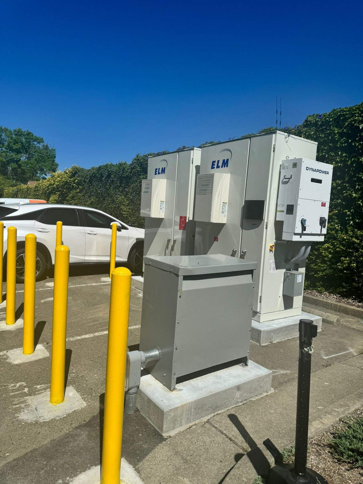 Pair of ELM battery energy storage cabinets with a Dynapower inverter installed on a concrete pad behind protective bollards in a Healdsburg, CA parking lot.