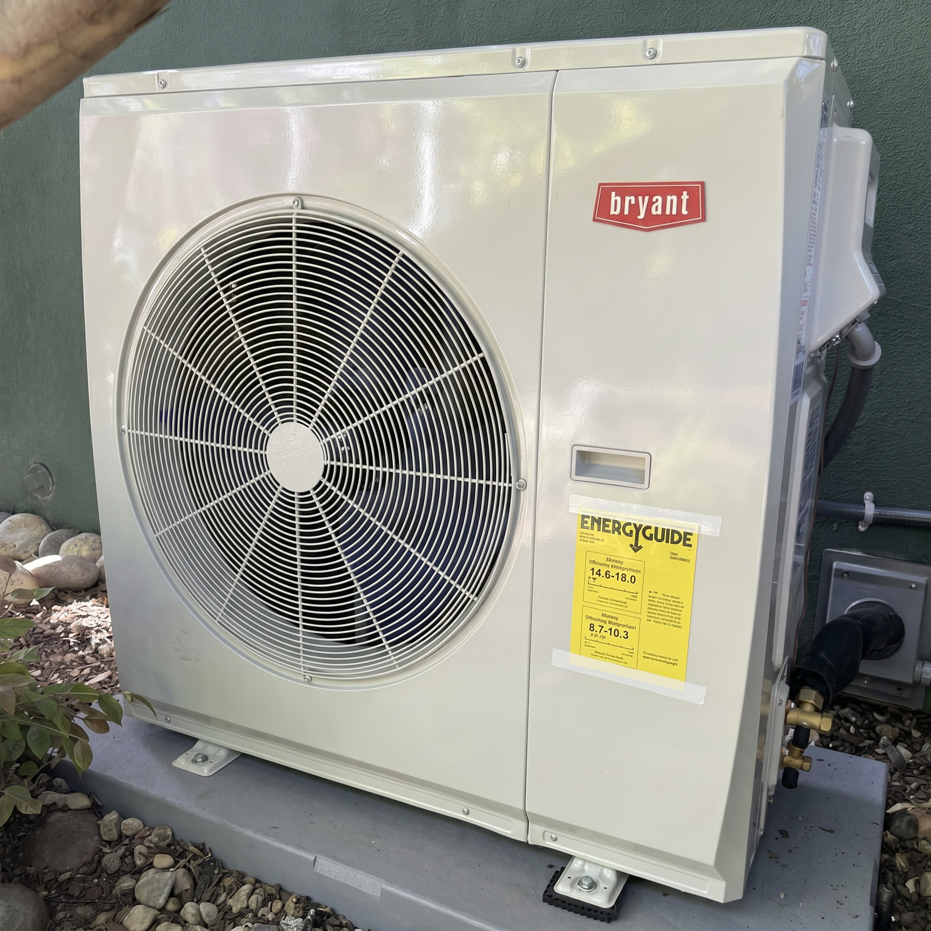 Newly installed white Bryant side-discharge heat pump condenser on a composite pad against a sage green stucco wall, with EnergyGuide sticker and service valves visible.