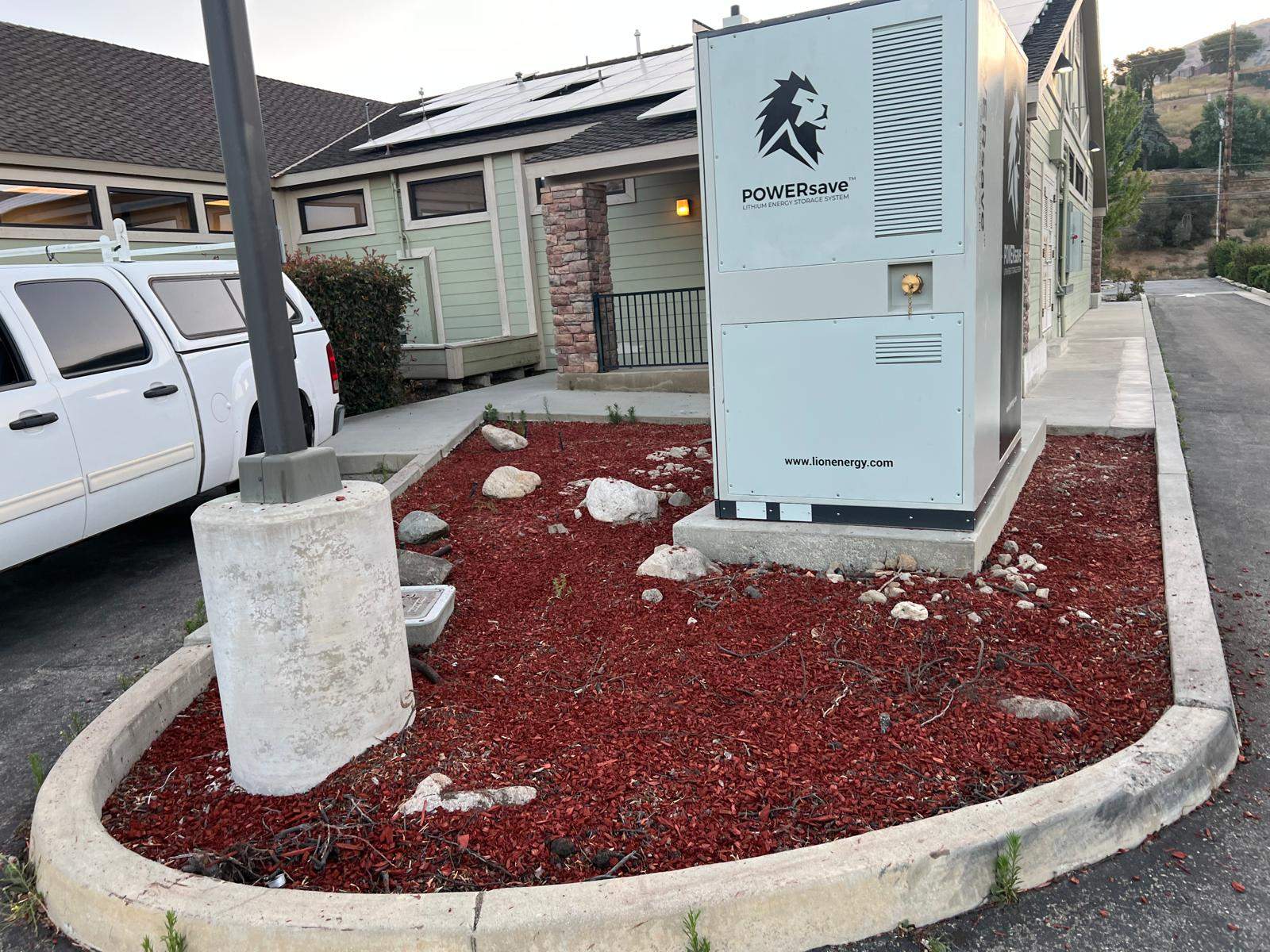 Lion Energy POWERsave lithium energy storage cabinet installed on a concrete pad in a red-mulch landscape bed outside a commercial building in Lebec, CA, with rooftop solar visible behind.