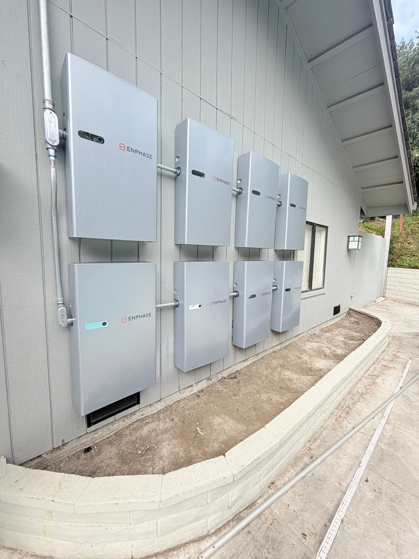 Eight Enphase IQ Batteries mounted in a two-by-four grid on the exterior wall of a Morgan Hill home, with conduit routed cleanly to a nearby subpanel.