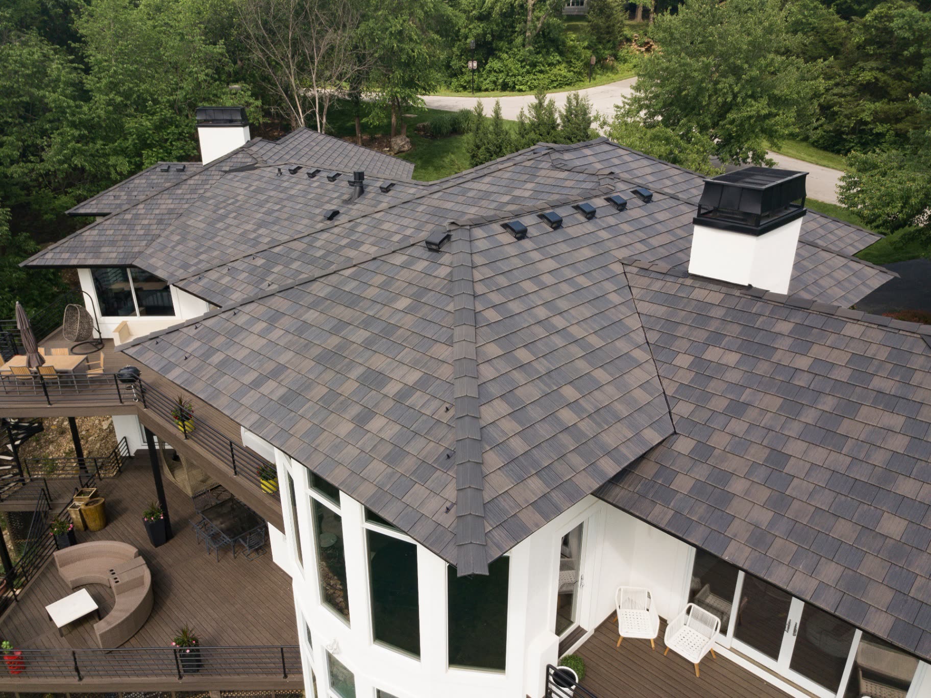 Aerial view of a contemporary home with a premium designer shingle roof in a blended black-oak finish, wrapped by mature trees and a wraparound deck.