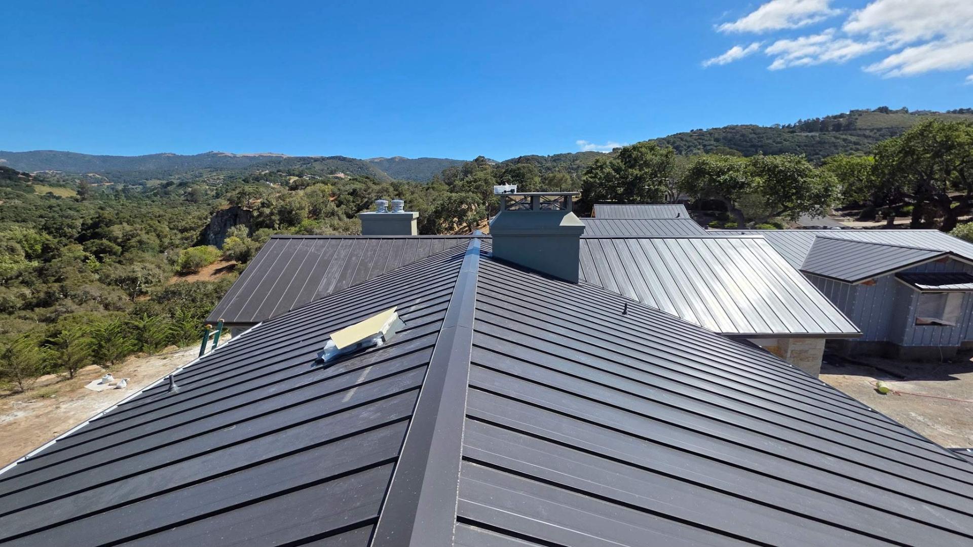 Matte-black standing-seam metal roof on a hillside California home with rolling oak-covered hills and blue sky in the background.