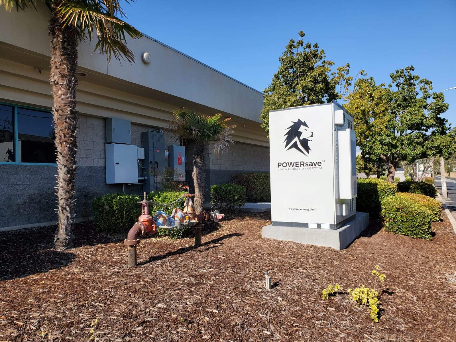 Lion Energy POWERsave lithium energy storage system installed on a concrete pad outside a commercial building in Santa Maria, CA, next to landscaping and utility service equipment.