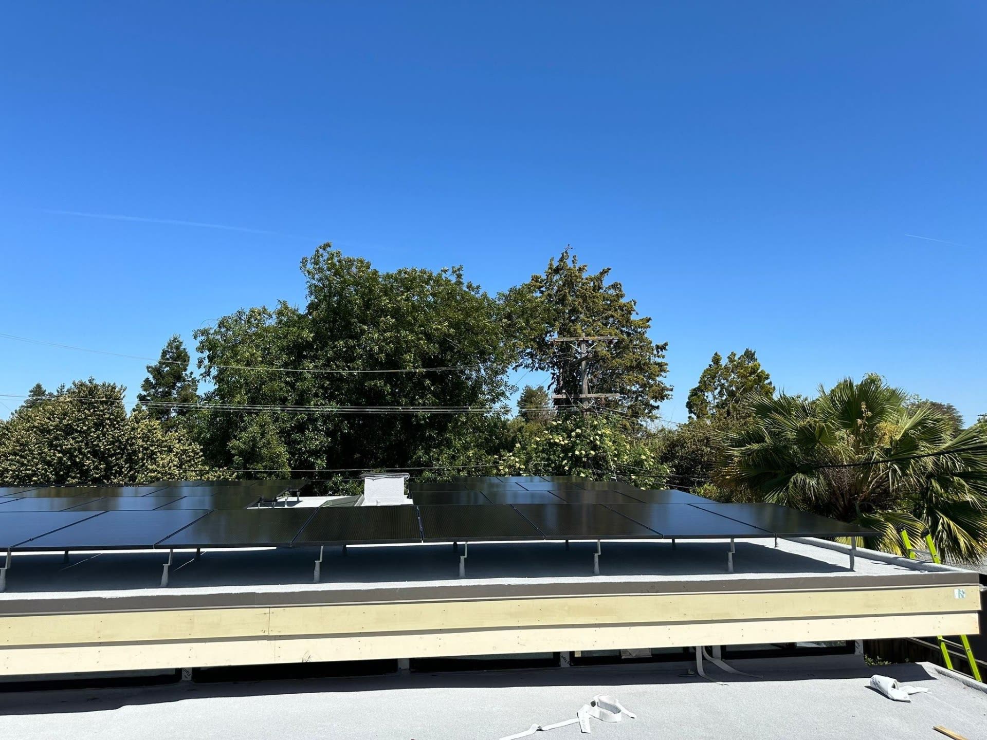 Tilt-racked all-black solar array elevated above a low-slope roof edge on a Sunnyvale home, framed by trees and blue sky.